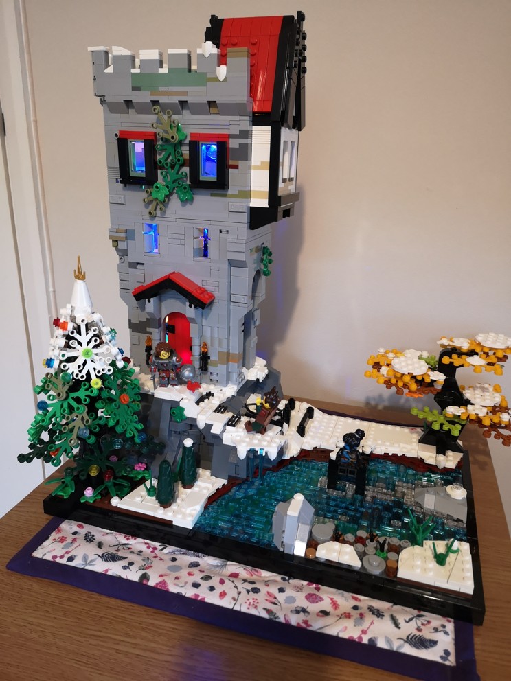 Lego diorama of a castle tower overlooking a bubbling river. The castle sits atop a rocky mound and appears to have been converted for habitation with the addition of a wooden extension clinging to its top. It is clearly winter, as the ground is covered in snow, save for the areas sheltered beneath the boughs of the trees, one of which has been decorated with baubles for the winter celebrations. Three figures are seen around the area. A tomboyish knight in full plate removed her helmet to greet her companion, a ranger with spiky hair and brown clothes. Sat by the river is a blue-skinned rogue, who appears to be the most feminine of the three.